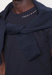 Tmavě modré bavlněné tričko s kulatým výstřihem a bílým logem "TOMMY HILFIGER". Přes ramena je přehozený tmavě modrý svetřík ve stejném odstínu.