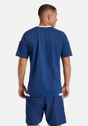 Mężczyzna odwrócony tyłem, ubrany w niebieską sportową koszulkę z krótkim rękawem i dopasowane niebieskie szorty na jednolitym białym tle.