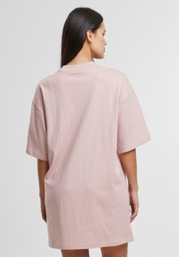 Preveliki ružičasti T-shirt haljina od mekanog materijala. Okrugli izrez, kratki rukavi i ležeran kroj, koji seže do sredine bedra s glatkom teksturom.