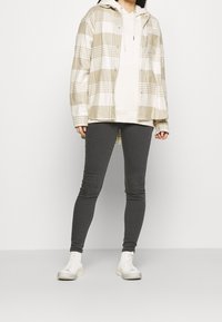 Giacca a scacchi beige e bianca sopra una felpa leggera, abbinata a leggings grigio scuro e scarpe high-top bianche.