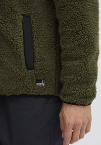 Olivenfarget fleece-genser med strukturert, luksuriøs overflate. Har en svart panel lomme og et lite merke som sier "11 PROJECT" nær kanten.