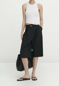 Person iført en hvid ærmeløs top, sorte knælangt brede shorts med bælte, sorte klipklapper, der holder en stor sort taske.