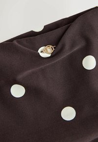 Tesătură de culoare maronie cu buline albe mari, având un sistem de închidere cu un buton ce are un accent circular de aur. Textură netedă, pliată pentru expunere.