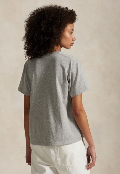 Žena s kučeravými vlasmi, oblečená v jednoduchom sivom tričku s krátkym rukávom a bielych nohaviciach, pozerá do strany na neutrálnom pozadí.