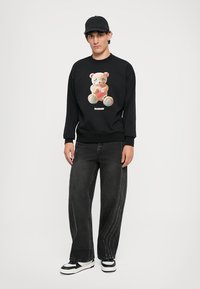 Felpa nera con grafica di un orsetto, abbinata a jeans scuri e sneakers nere. Il modello indossa un cappellino nero.