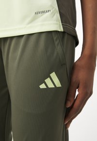 Ljust grön och mörkgrå atletisk tröja med en strukturerad design; olivgröna byxor med en reflekterande Adidas-logotyp och dragsko i midjan.