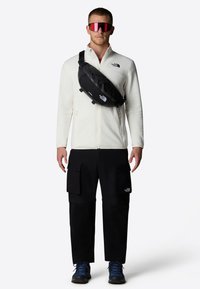 Bolso bandolera negro con logo, llevado sobre una chaqueta de forro polar blanca y pantalones cargo negros. Presenta grandes bolsillos, tela resistente y zapatos de senderismo azules.