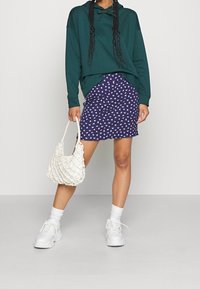 Grön hoodie, lila kjol med vitt fjärilsmönster, vita sneakers och textured vit väska. Modellen står mot en neutral bakgrund.