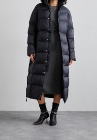 Černý péřový kabát s dlouhým prošitým designem, zapínáním na patentky, bočními rozparky, nošený přes žebrované šedé šaty, doplněný černými kotníkovými botami.