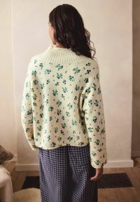 Hele kollane kardigan, millel on roheline lillemuster, soonikkrae ja lühike siluett. Kandmine siniste ja valgete ruuduliste pükste juurde.