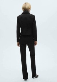 Costume noir ajusté avec une veste cintrée et un pantalon fuselé. Le tissu a l'air lisse, et la veste a un col structuré.