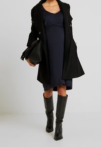Marinblå klänning med samlad detaljer, lager under en svart kappa. Kombinerad med knähöga, spetsiga svarta läderstövlar. Stor svart handväska.