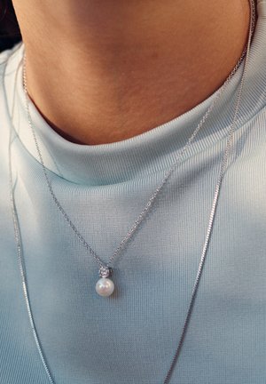 Silver necklace featuring a round pearl pendant, accompanied by a small, clear stone, worn over a light blue textured shirt.