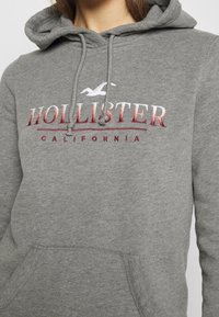 Grå hoodies med en framficka, snörhuva och broderad "Hollister California"-logga i vitt och röd gradient. Mjuk material, avslappnad passform.