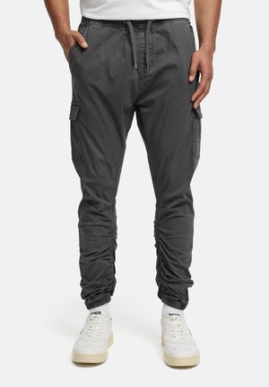 Homme portant un pantalon de jogging cargo gris foncé avec des poignets froncés et des baskets blanches, les mains dans les poches, debout devant un fond uni.