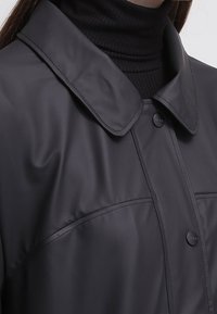 Svart vattentät jacka med uppslagbar krage, frontknäppning och slät textur. Diskreta sömdetaljer över bröstet.