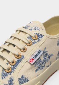 Beige lerretsneaker med snøring, blått naturskjønt trykk, gullfargede nagler og en hvit merkelapp med "Superga" på siden.