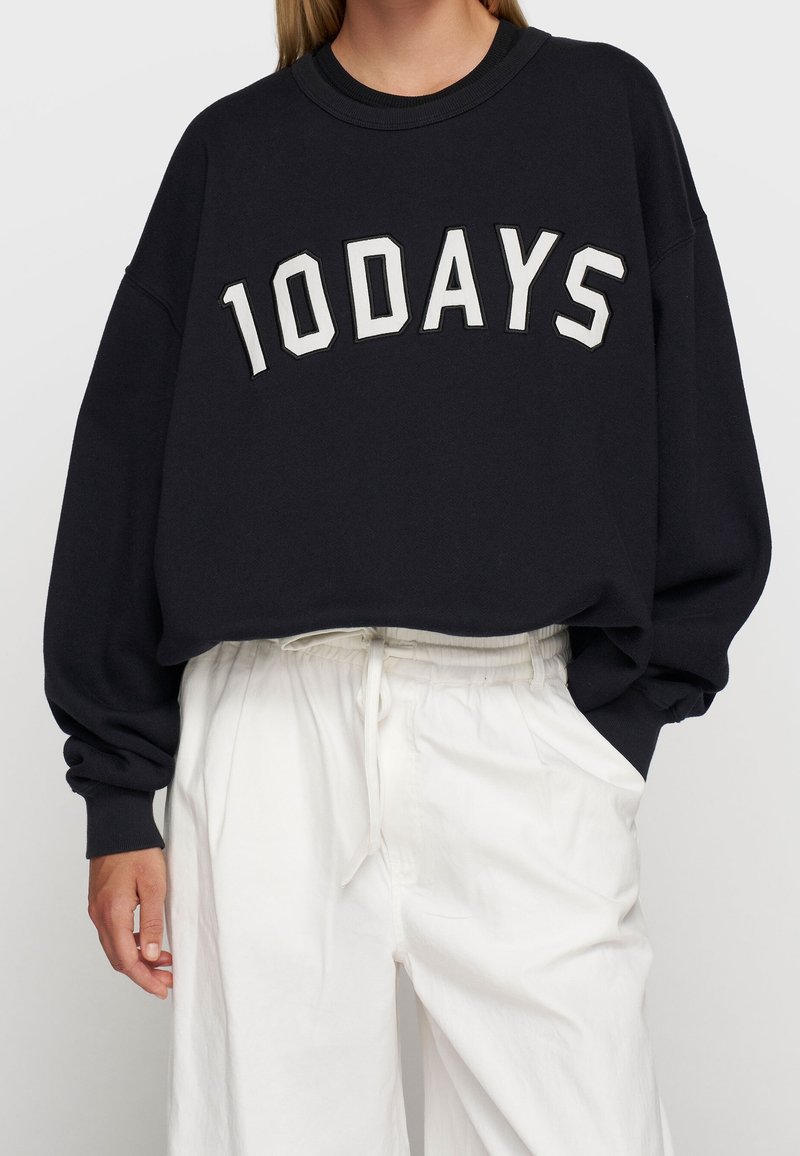 Osoba ubrana w luźną czarną bluzę z napisem "10 DAYS" oraz białe spodnie z trokami, z ręką swobodnie włożoną do kieszeni spodni.
