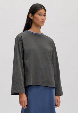 Jonge vrouw met lang donker haar die een loszittende donkergrijze top met lange mouwen draagt over een blauwe rok tegen een effen lichte achtergrond.