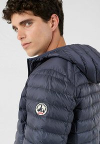 Doudoune marine avec capuche, présentant un design matelassé, une texture légère et un patch circulaire avec logo sur l'épaule gauche.