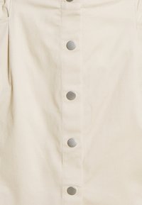 Beige kjol med en slät yta, som har sex metalldragknappar ned längs mitten och subtila veck vid midjan.