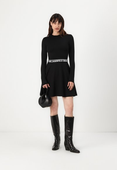 Robe noire à manches longues avec jupe évasée, ceinture de marque blanche, associée à des bottes noires brillantes montantes et un petit sac à main noir.