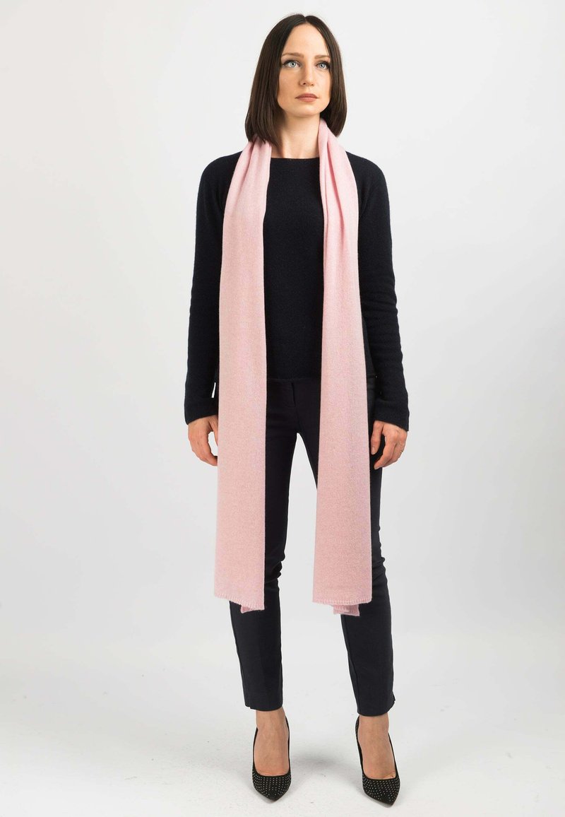 Sciarpa rosa chiaro in morbido materiale, drappata liberamente attorno al collo. Indossata con un maglione nero e pantaloni scuri. Scarpe con tacchi alti completano il look.