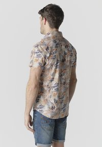 Hombre de espaldas que lleva una camisa de manga corta con estampado floral de hojas beige, naranja y moradas, combinada con pantalones cortos vaqueros remangados.