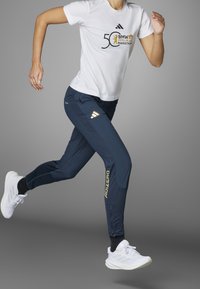 Adidaksen juoksuasu, jossa on valkoinen t-paita mustalla ja kultaisella logolla sekä laivastonsiniset housut, joissa on kultaiset "ADIZERO"-kirjaimet ja sivuraidat.