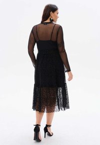 Czarna sukienka z przezroczystą, długą koronką i warstwową spódnicą w panterkę. Zasłonięty tył z detalem guzikowym. Czarne szpilki.
