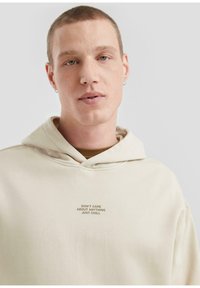 Primo piano di una persona con capelli corti che indossa un hoodie beige chiaro con la scritta "NON MI INTERESSA NULLA, RILASSATI E BASTA" sul petto.