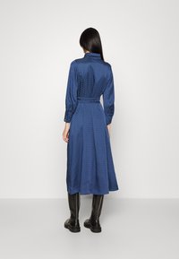 Lång blå klänning i texturerad tyg, med bälte, knappade manschetter och krage. Bärs med svarta stövlar som går till mitten av vadskalet.