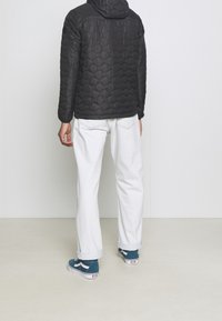 Giacca a vento nera trapuntata con cappuccio, jeans bianchi a gamba dritta e sneakers color teal. Texture notevoli e colori a contrasto.