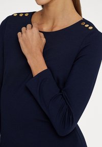 Bluză cu mânecă lungă de culoare bleumarin, având un guler rotund, trei detalii cu nasturi aurii pe umăr și un material moale, texturat.