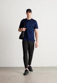 Homem a caminhar, vestindo uma t-shirt azul-marinho, boné preto, calças pretas ajustadas, sapatilhas pretas e a segurar uma bolsa de ombro preta.