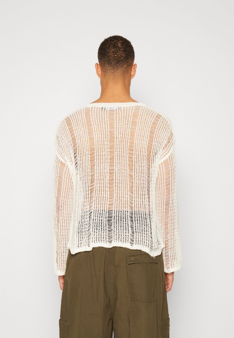 Man som vänder bort ansiktet, iklädd en vit löst stickad långärmad tröja som visar huden under och olivgröna cargo-byxor.