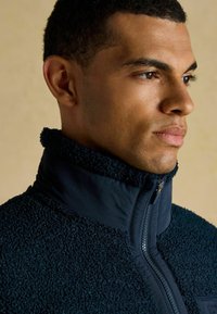 Mies, jolla on lyhyet kiharat hiukset, pukeutuneena tummansineään fleece-takkiin, jossa on korkea vetoketjukaulus, katsoo oikealle yksinkertaista taustaa vasten.