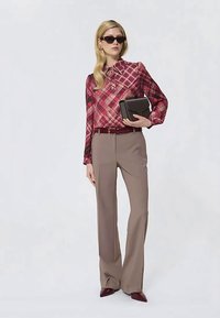Femme portant une blouse à carreaux rouges, un pantalon large beige, des chaussures pointues bordeaux, des lunettes de soleil, tenant un sac à main noir.