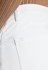 Close-up van een persoon die een witte broek draagt met een gestikte hartdetail op de achterzak en een bruine geribbelde trui die is ingestopt.