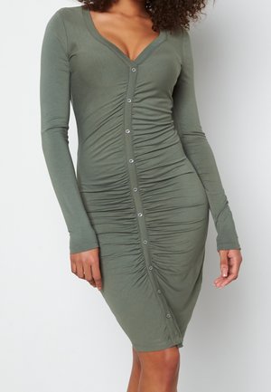 Frau trägt ein eng anliegendes olivgrünes Kleid mit langen Ärmeln, vorne mit Knöpfen und gerafften Details in der Mitte.