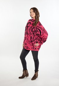 Übergroßer rosa Pullover mit schwarzen abstrakten Mustern, Rundhalsausschnitt, gerippte Ärmelbündchen, getragen mit dunklen Jeans und braunen Stiefeletten.