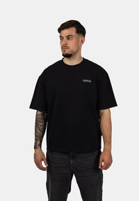Černé bavlněné tričko s krátkými rukávy, volný střih, kulatý výstřih a malý bílý text "CRYSTAL" na levé části hrudi.