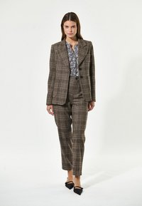 Blazer a quadri marrone con pantaloni abbinati in tessuto testurizzato. Il blazer presenta ampi risvolti e due bottoni. Scarpe ballerine nere a punta.