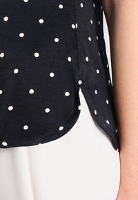 Blusa blu navy con pois bianchi, realizzata in tessuto leggero. Presenta un orlo curvo e spacchi laterali per facilitare i movimenti.