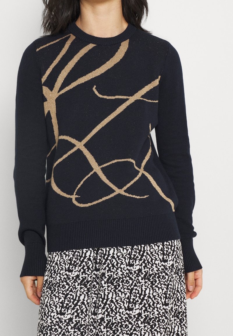 Pull bleu marine avec un motif abstrait métallique doré. Col rond, poignets et ourlet côtelés. Associé à une jupe à motifs noirs et blancs.