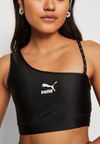 Crop top nero con spallina unica, realizzato in un materiale liscio e con il logo Puma bianco. Presenta una tracolla decorata con piccoli loghi Puma.