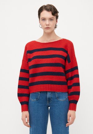 Kvinne iført en rød genser med svarte horisontale striper og blå jeans, som står mot en ensfarget hvit bakgrunn.