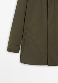 Lang olijfgroene jas met een rechte snit. Voorzien van een enkele borstzak, opgevouwen manchetten en een gladde, matte textuur.