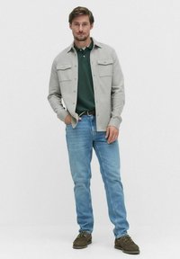 Hellgraues Hemd mit Knopfleiste und zwei Brusttaschen über einem dunkelgrünen Poloshirt, kombiniert mit blauen Jeans und braunen Wildlederschuhen.