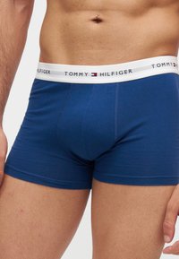 Мъжки торс, облечен в сини боксерки с бял колан с марка Tommy Hilfiger, показващ пиърсинг на пъпа.
